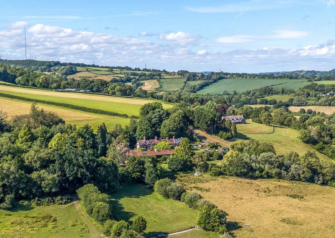 Halshayne Manor Farm seen from the air in summer, its buildings set among trees in a green valley of fields and hedgerows.