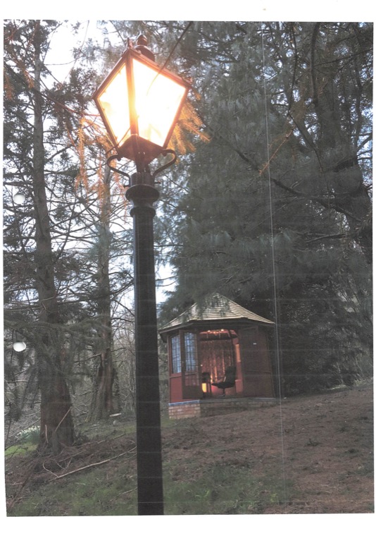 The summer house at dusk among conifers, glowing from within, with a lit Victorian standard lamp in the foreground.